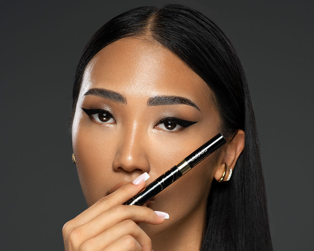 Close up of a woman holding Qudos eyeliner pen with lid just below her left eye, grey background
