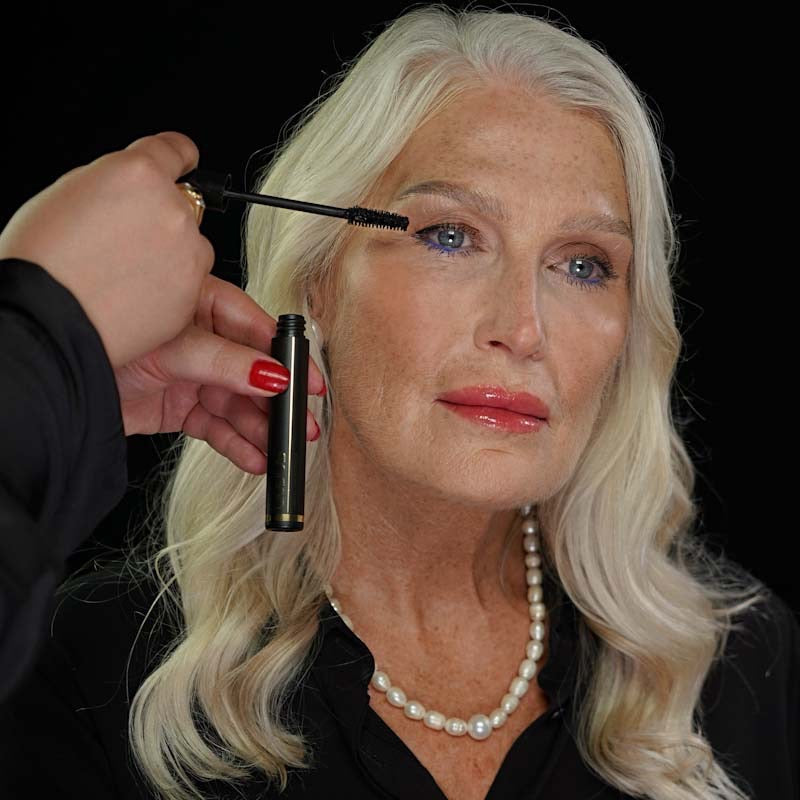 Close up older woman having Qudos I Wand You mascara applied to her right eye by somebody else, black background