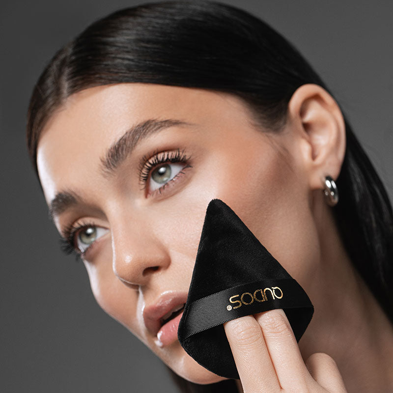 Woman wearing natural face makeup with mascara looks away from camera while holding synergy powder puff against her left cheek