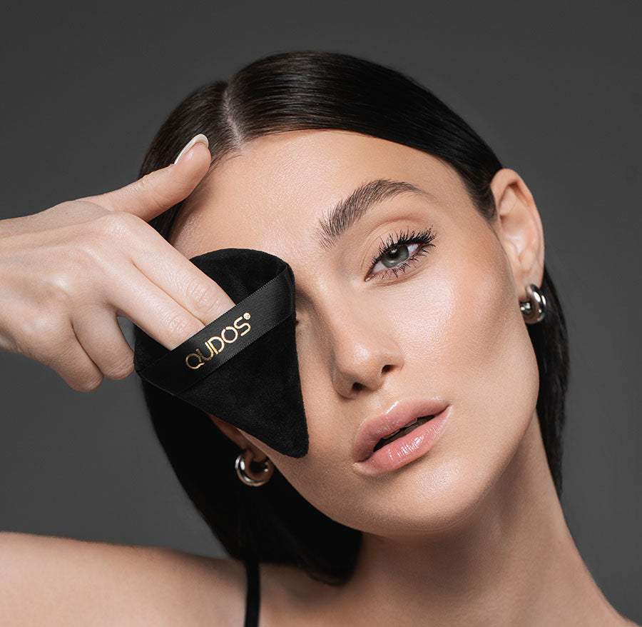 Woman looking into camera while holding Synergy powder puff in front of her right eye with Qudos name on display