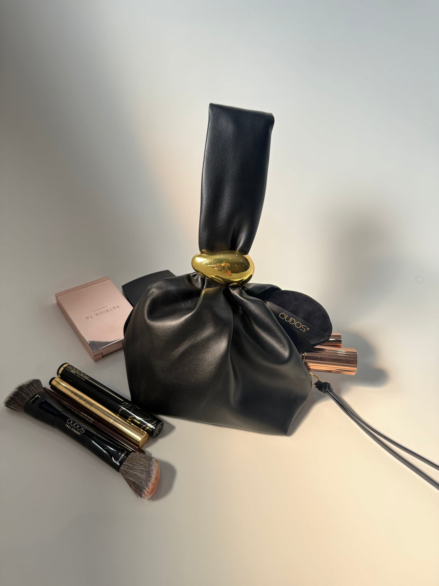 Qudos black drawstring luxury makeup bag with makeup brushes and products on a gradient background