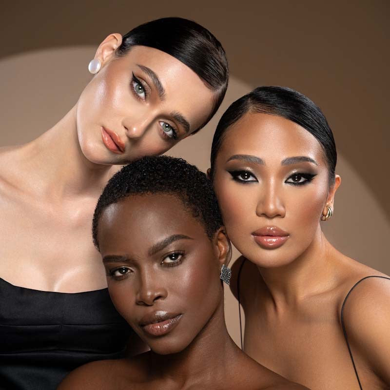 Three women of different skin tones with natural face makeup posing and looking into camera.
