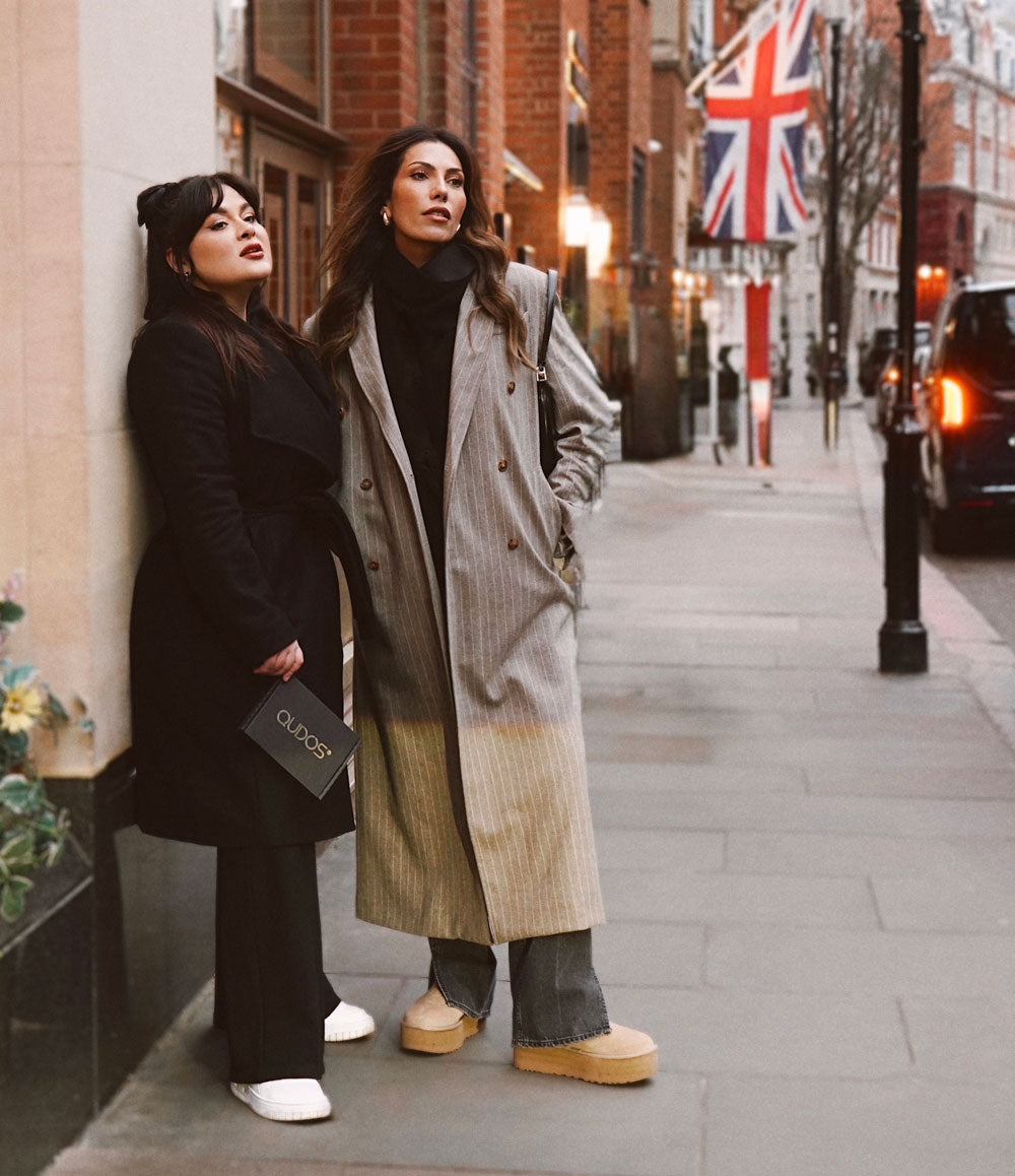 Company directors Andra and Ana-Maria stood by the wall of a street looking away from the camera, out of focus taxi and buildings in the background 