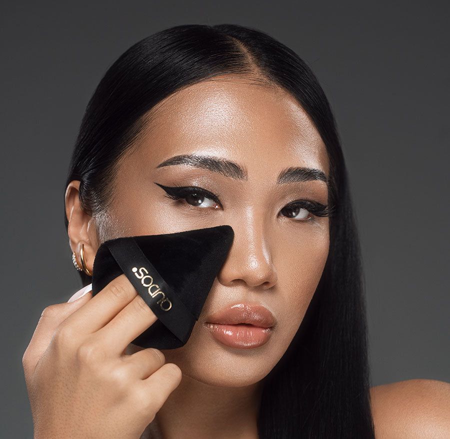 Asian woman with natural face makeup and winged eyeliner holding Synergy powder puff below right eye while looking into camera