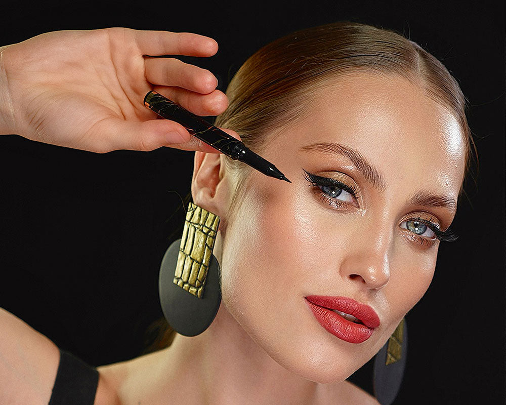 Close up of woman staring into camera with Qudos eyeliner pen held next to her right eye 